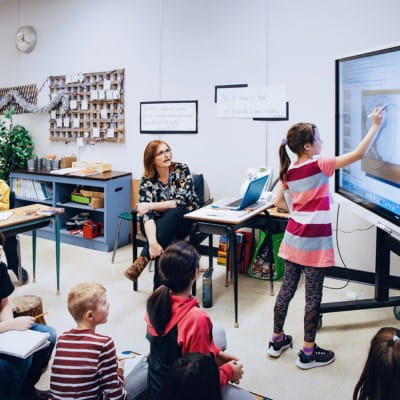 A student engaging with content on a SMART interactive display, while her teacher and classmates watch and support.