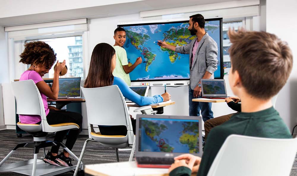 Interactive classroom with students using laptops and a teacher pointing at a SMART board displaying a world map, indicative of a geography lesson in a technologically equipped educational environment.