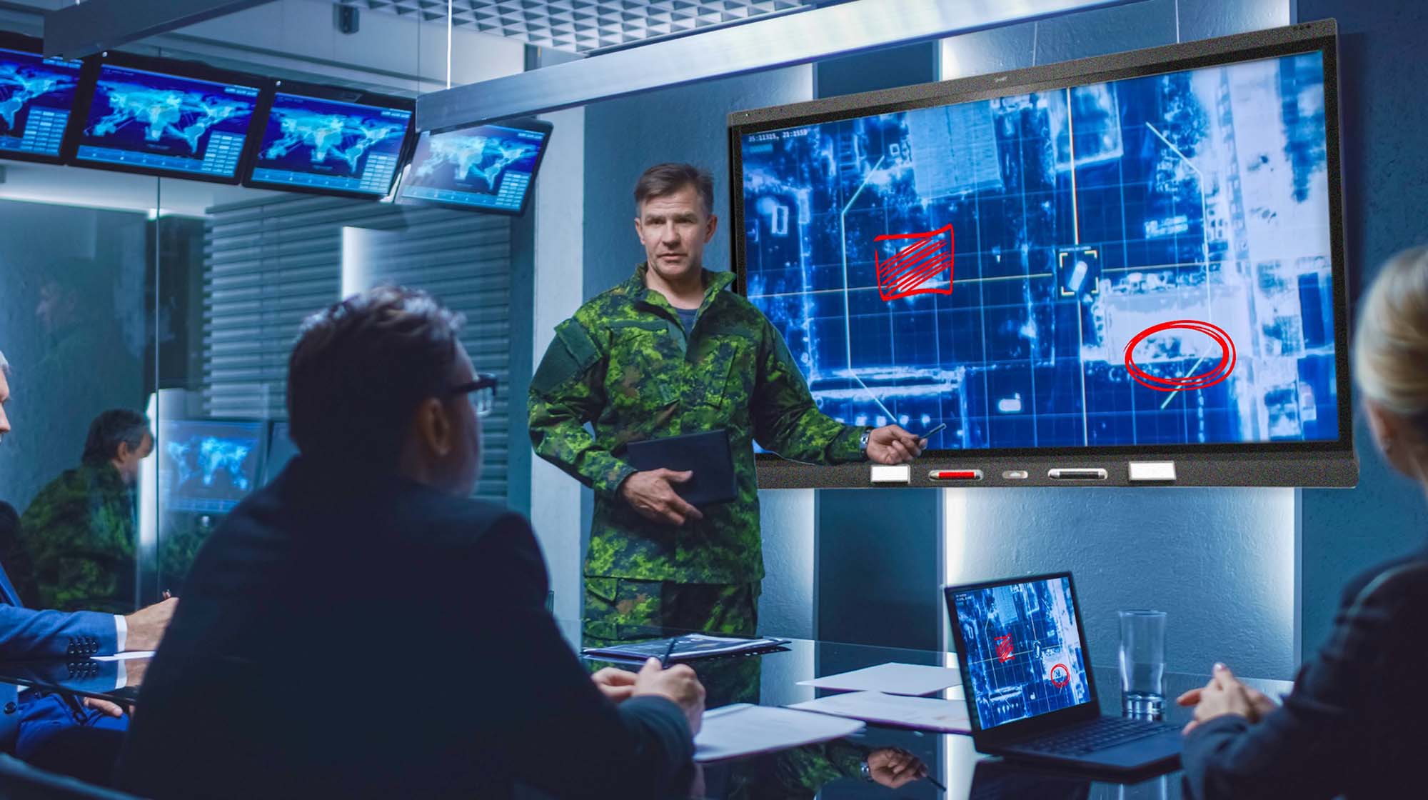 Military officer giving a presentation using the SMART board with marked satellite images in a high-tech briefing room.