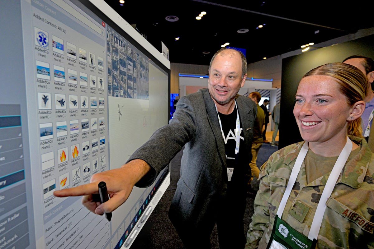 A business professional demonstrating the capabilities of an interactive SMART board to a smiling Air Force officer at a tech conference.