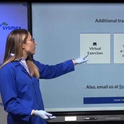 Woman in blue lab coat and gloves pointing at a SMART board displaying 'Virtual Exercises' and other training options, with a Sysmex logo at the top.
