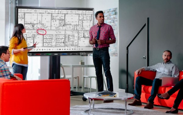 A man stands presenting at a SMART Board interactive display in a modern office space, with architectural plans on screen, indicating active collaboration.